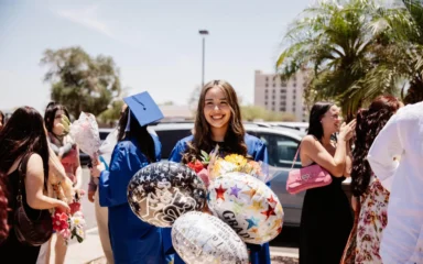 Genesis, wearing a blue graduation gown and holding balloons and a bouquet of flowers, smiles brightly while celebrating her graduation outside with friends and family.