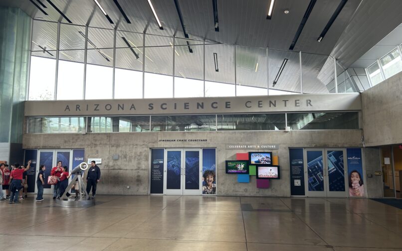 Lobby of the Arizona Science Center with large windows, concrete walls, and displays along the back wall; a group of visitors stands on the left near an exhibit