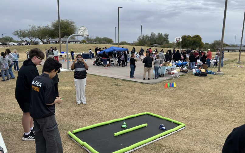 Outdoor event at a park with groups of people gathered around tables and activity stations near a basketball court. In the foreground, two individuals stand near a small foam-bordered racetrack on the grass, with toy cars positioned inside it. A person in the mid-ground is taking a photo, and tents, cones, and various activity setups are visible throughout the area under cloudy skies.