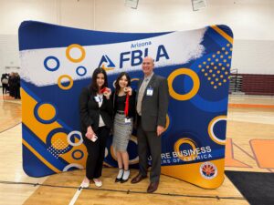 Three people stand in front of a large blue, white, and orange Arizona FBLA backdrop at an indoor gymnasium event. The person in the center holds up a medal with a red ribbon. All three are wearing business attire and event name badges. Other attendees can be seen in the background on the left side of the gym.