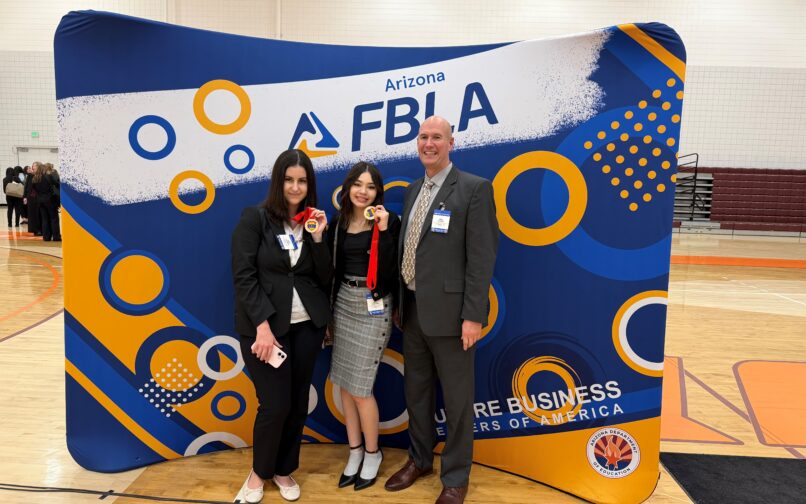 Three people stand in front of a large blue, white, and orange Arizona FBLA backdrop at an indoor gymnasium event. The person in the center holds up a medal with a red ribbon. All three are wearing business attire and event name badges. Other attendees can be seen in the background on the left side of the gym.