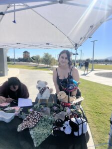 A woman smiles beside a table displaying dog accessories under a gazebo at a park.