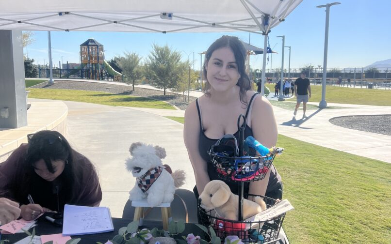 A woman smiles beside a table displaying dog accessories under a gazebo at a park.